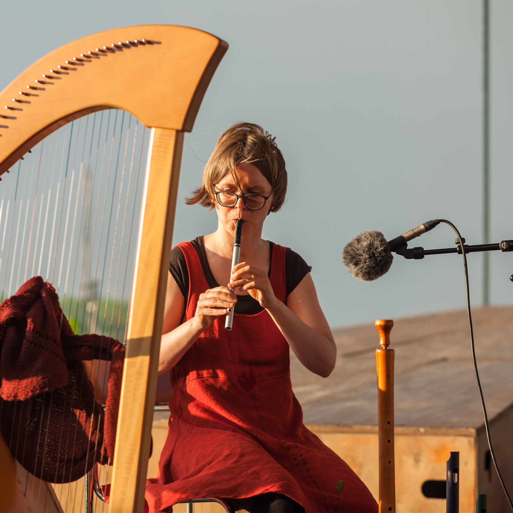 Katharina Hölzel spielt im Wind auf einer irischen Whistle. Neben ihr steht eine Hakenharfe deren Saiten von einem Tuch schützend durchwoben sind.