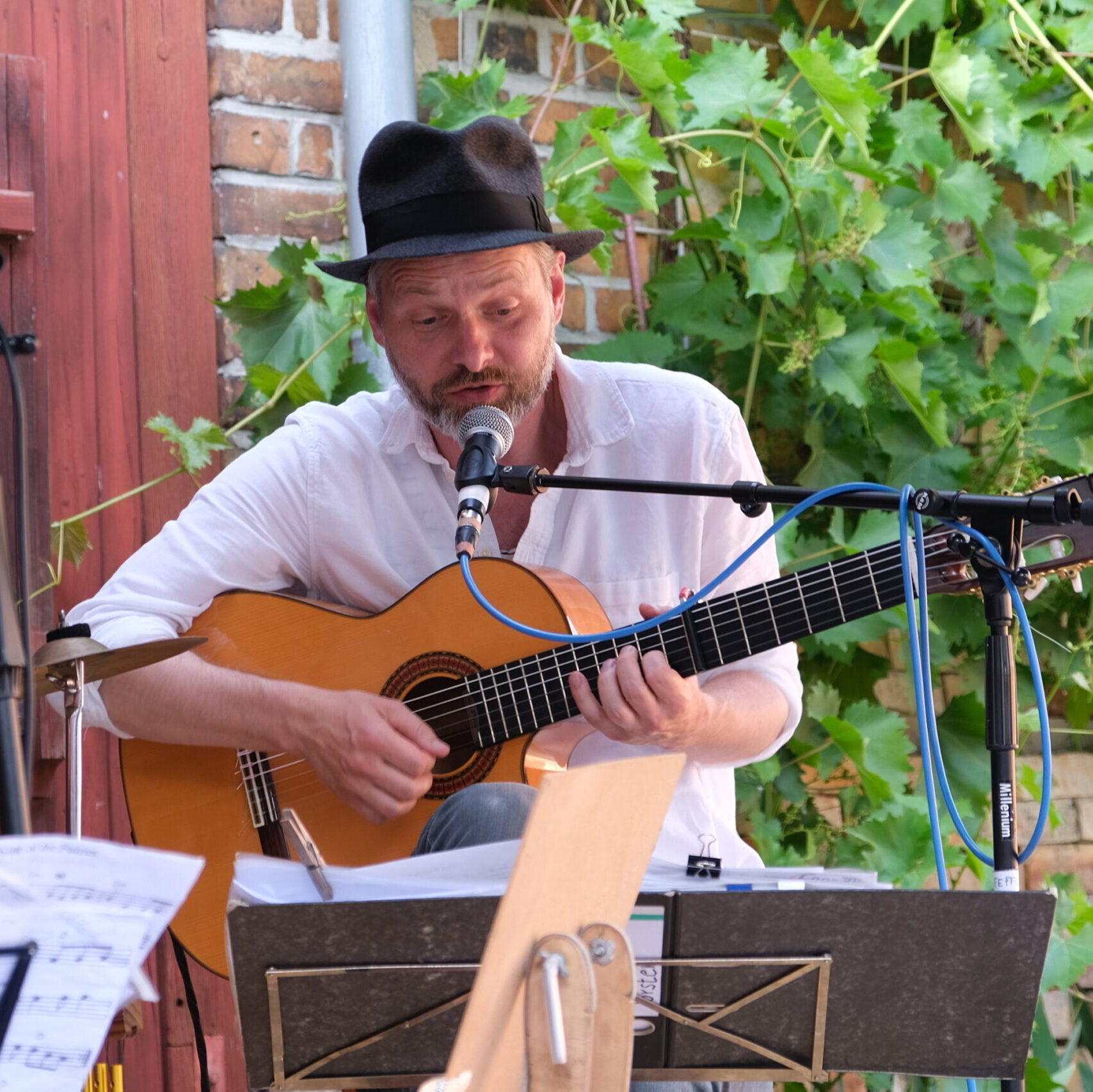 Der Gitarrist Torsten Pfeffer sitzt vor einer Weinbewachsenen Backsteinmauer während er seinen Gesang auf der Gitarre begleitet. Er trägt Hut und Hemd. Im Hintergrund zeichnet sich das Tor eines Bauernhofs ab.